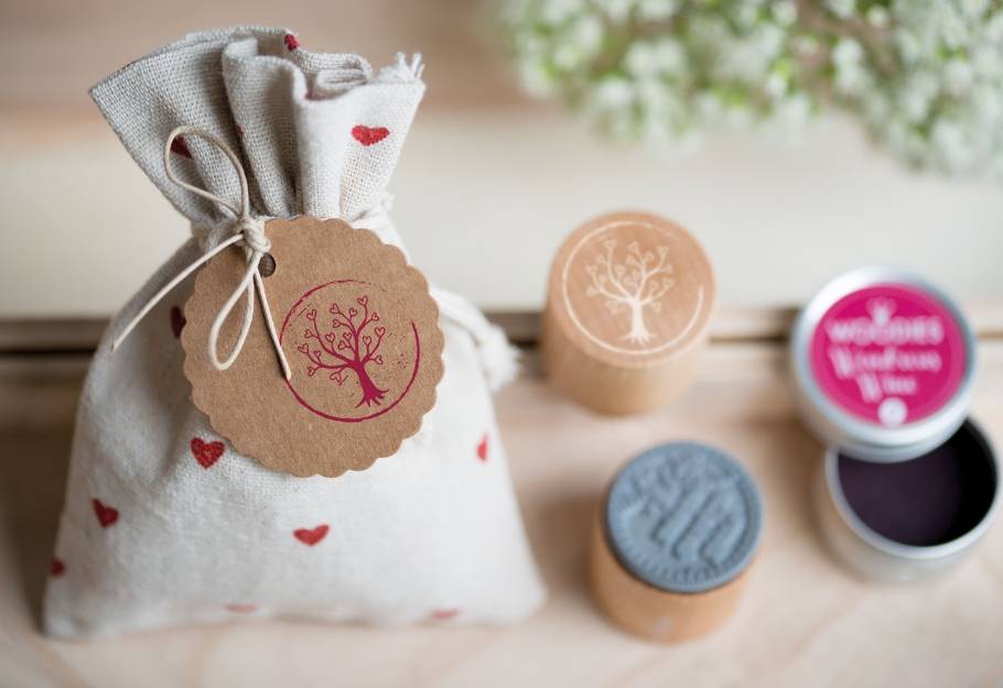 Ein Leinenbeutel mit aufgedruckten roten Herzen und einem runden, braunen Anhänger, auf dem ein Baum in rot gestempelt ist, steht auf einem Holztisch. Daneben befindet sich ein Holzstempel mit dem gleichen Baummotiv und ein geöffnetes Stempelkissen mit der Aufschrift 